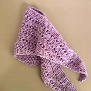 May include: A lilac-coloured, hand-knitted shawl with a triangular shape. The shawl has a delicate openwork pattern throughout the fabric. The ends of the shawl are pointed, and the fabric appears soft and lightweight. The shawl is displayed on a neutral background.