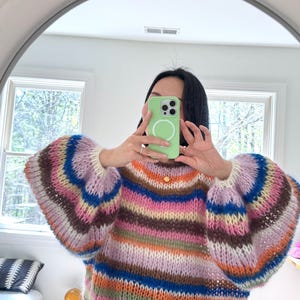 Può includere: Un maglione oversize colorato con righe orizzontali nei toni del marrone, rosa, blu e arancione. Il maglione ha maniche ampie e a sbuffo e uno scollo tondo. L'immagine è scattata davanti a uno specchio.