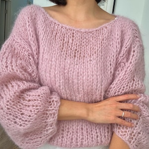 Op de afbeelding: Een lichtroze, open gebreide trui met een brede halslijn en lange, volumineuze mouwen. De trui heeft een iets kortere pasvorm en een zachte, fuzzy textuur. Het kledingstuk is gemaakt van een delicaat materiaal, mogelijk mohair of iets dergelijks.