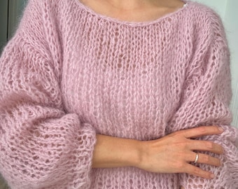 May include: A light pink, open-knit sweater with a wide neckline and long, voluminous sleeves. The sweater has a slightly cropped fit and a soft, fuzzy texture. The garment is made of a delicate, possibly mohair or similar, material.