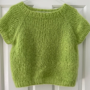 May include: A lime green, short-sleeved sweater with a round neckline. The knitted top is hanging on a silver hanger against a white background. The sweater's texture is visible, showcasing the yarn's weave.