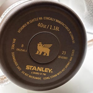 May include: Close-up of the bottom of a brown Stanley brand water bottle. The bottle is marked "40oz / 1.18L" and "Dishwasher Safe". The Stanley logo is visible, along with other text.