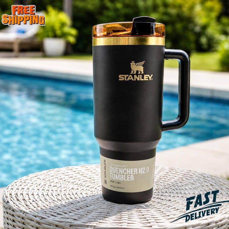 May include: Black Stanley Quencher H2.0 Tumbler with a gold rim and handle. The tumbler has the Stanley logo and the text "QUENCHER H2.0 TUMBLER" on the side. It is sitting on a white woven table near a pool.