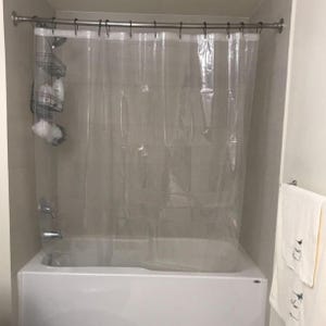 May include: A clear, transparent shower curtain hangs in front of a white bathtub. A metal shower rod and hooks hold the curtain. A metal caddy with toiletries and a loofah is mounted on the wall. White towels hang on a towel rack.
