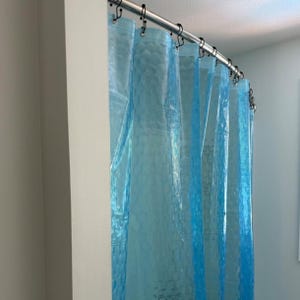 May include: A blue shower curtain with a textured pattern hangs from a curved metal rod with black rings. The curtain is made of a translucent material, allowing light to pass through. The shower curtain is in a bathroom setting.
