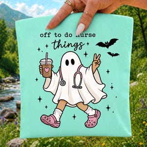 May include: A light mint green tote bag featuring a graphic of a white ghost wearing a pink stethoscope and pink clogs. The ghost holds a coffee cup with a heart icon and makes a peace sign. The text reads off to do nurse things with bats.