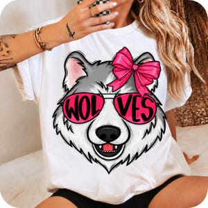 Peut inclure: Un t-shirt blanc à manches courtes orné d'un motif représentant une tête de loup gris portant des lunettes de soleil rose vif avec le texte WOLVES imprimé sur les verres. Le loup porte un grand nœud rose vif sur le dessus de la tête.
