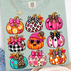 Può includere: Una t-shirt verde salvia con una stampa grafica di nove zucche stilizzate. Le zucche sono decorate con vari motivi, tra cui stampa leopardata, quadri, pois e disegni floreali, accentuati da fiocchi, cuori e stelle.