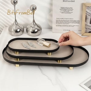 Puede incluir: Bandejas ovaladas de tres niveles con bordes negros e interiores beige, sostenidas por patas doradas. Las bandejas contienen joyas y un lápiz labial. Dos candelabros plateados están en el fondo. Las bandejas están sobre una superficie blanca.
