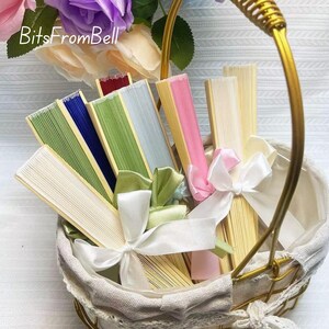 May include: A collection of colorful folding fans in a white basket with a gold handle. The fans are in various colors, including green, blue, pink, and cream, and are tied with white and green ribbons. The text "BitsFromBell" is visible.