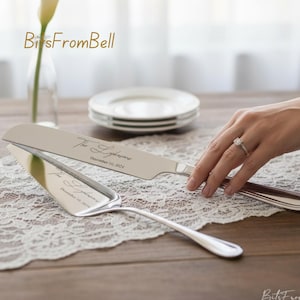 May include: Silver cake knife and server set with engraved text. The knife and server are displayed on a lace tablecloth. The text reads "The Lydersons" and "December 10, 2024". A hand with a ring is holding the knife.