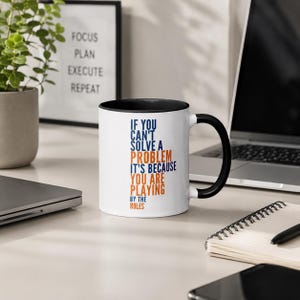 May include: White ceramic mug with a black interior and handle. The mug features the text "IF YOU CAN'T SOLVE A PROBLEM IT'S BECAUSE YOU ARE PLAYING BY THE RULES" in blue and orange. A motivational office or home decor item.