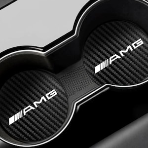 May include: Black car cup holder inserts with a carbon fiber pattern and the white letters "AMG". The inserts are round and fit into the car's cup holders. The car's interior is black and gray.