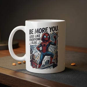 K&ouml;nnte beinhalten: Wei&szlig;e Keramik-Tasse mit einer Comic-Illustration von Spider-Man und dem Text "BE MORE YOU. LESS LIKE EVERYONE ELSE." Der Superheld ist in einem rot-blauen Anzug auf einem Dach dargestellt. Die Tasse hat einen gebogenen Griff.