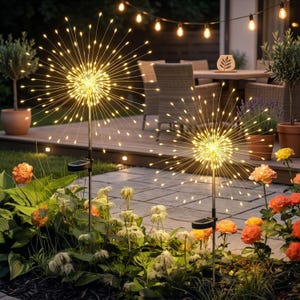 Op de afbeelding: Twee tuinlampen op zonne-energie met een vuurwerkachtig ontwerp. Elke lamp heeft een centrale bol met talrijke uitstralende LED-strengen, die een heldere, warme gloed creëren. De lampen staan in een tuin met bloemen en een terras op de achtergrond.