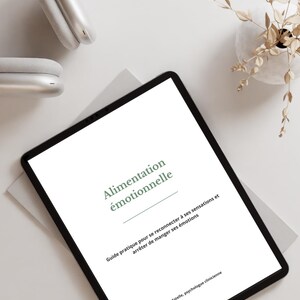 Peut inclure: Une tablette numérique à cadre noir affiche le titre "Alimentation émotionnelle" en vert. La tablette est posée sur une surface blanche, avec un casque blanc et un petit vase avec des plantes séchées à proximité. Le texte en dessous du titre se lit comme "Guide pratique pour se reconnecter à ses sensations et arrêter de manger ses émotions."