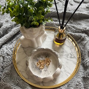 Könnte beinhalten: Ein rundes Tablett mit goldenem Rand und weißer Marmoroberfläche enthält einen weißen büstenförmigen Übertopf mit einer grünen Pflanze, einen Glas-Diffusor mit schwarzen Stäbchen und eine weiße, gewellte Harzschale mit zwei goldenen Ohrringen.