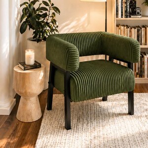 May include: A cozy reading nook featuring a green corduroy armchair with black legs, a wooden side table, and a bookshelf filled with books. A framed print with the text "Henri Matisse" hangs on the wall, and a floor lamp provides soft lighting.