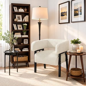 Puede incluir: Un sal&oacute;n acogedor con un sill&oacute;n de pana crema con patas de metal negro. Una estanter&iacute;a de madera oscura, mesas auxiliares y una l&aacute;mpara de pie con pantalla beige completan el espacio. Dos cuadros de paisajes enmarcados cuelgan de la pared.