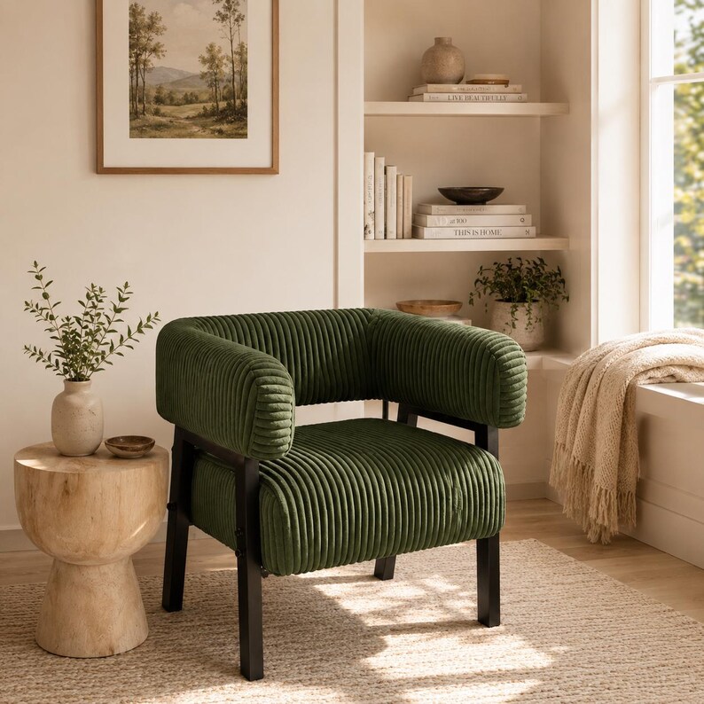 Puede incluir: Un sill&oacute;n de pana verde con estructura de metal negro. La silla tiene reposabrazos redondeados y una textura acanalada. La habitaci&oacute;n incluye una mesa auxiliar de madera y una pintura de paisaje enmarcada.