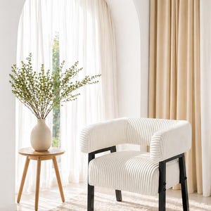 Puede incluir: Sill&oacute;n de pana color crema con estructura de metal negro. La silla tiene respaldo y reposabrazos redondeados. Una peque&ntilde;a mesa auxiliar de madera con un jarr&oacute;n con vegetaci&oacute;n est&aacute; cerca. La habitaci&oacute;n tiene cortinas neutras y una alfombra.