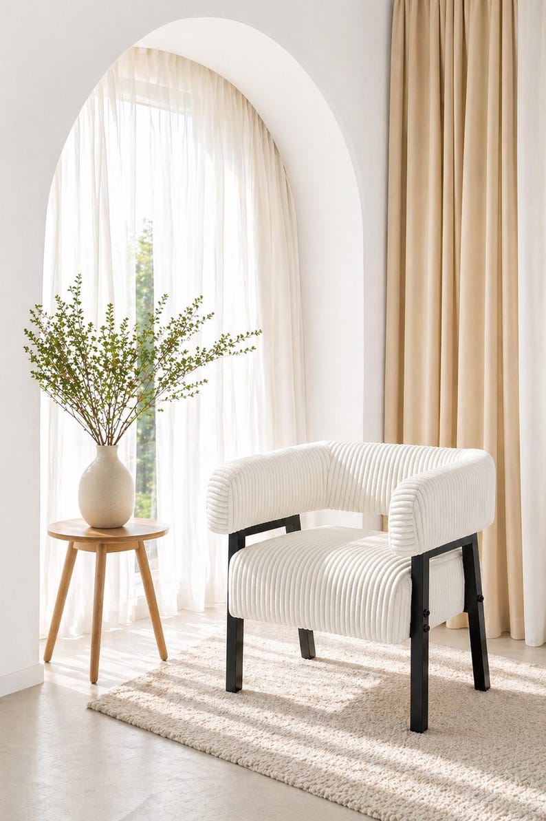 Puede incluir: Sill&oacute;n de pana color crema con estructura de metal negro. La silla tiene respaldo y reposabrazos redondeados. Una peque&ntilde;a mesa auxiliar de madera con un jarr&oacute;n con vegetaci&oacute;n est&aacute; cerca. La habitaci&oacute;n tiene cortinas neutras y una alfombra.