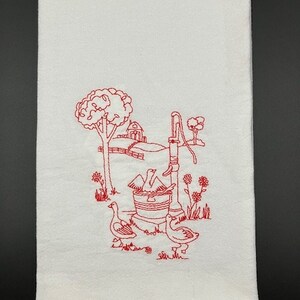 May include: A white kitchen towel with red embroidery of a farmhouse scene. The scene includes a well with a bucket, a small house, trees, and three birds.