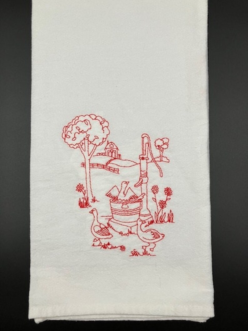 May include: A white kitchen towel with red embroidery of a farmhouse scene. The scene includes a well with a bucket, a small house, trees, and three birds.
