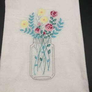 Puede incluir: Una toalla de cocina blanca con un diseño bordado de un tarro de cristal lleno de flores rosas y amarillas.