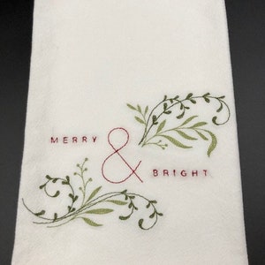 Puede incluir: Toalla de mano blanca con texto bordado y dise&ntilde;o bot&aacute;nico. Las palabras "MERRY & BRIGHT" est&aacute;n bordadas con hilo rojo. Hojas y vides verdes y oliva est&aacute;n bordadas alrededor del texto, creando un tema festivo de vacaciones.