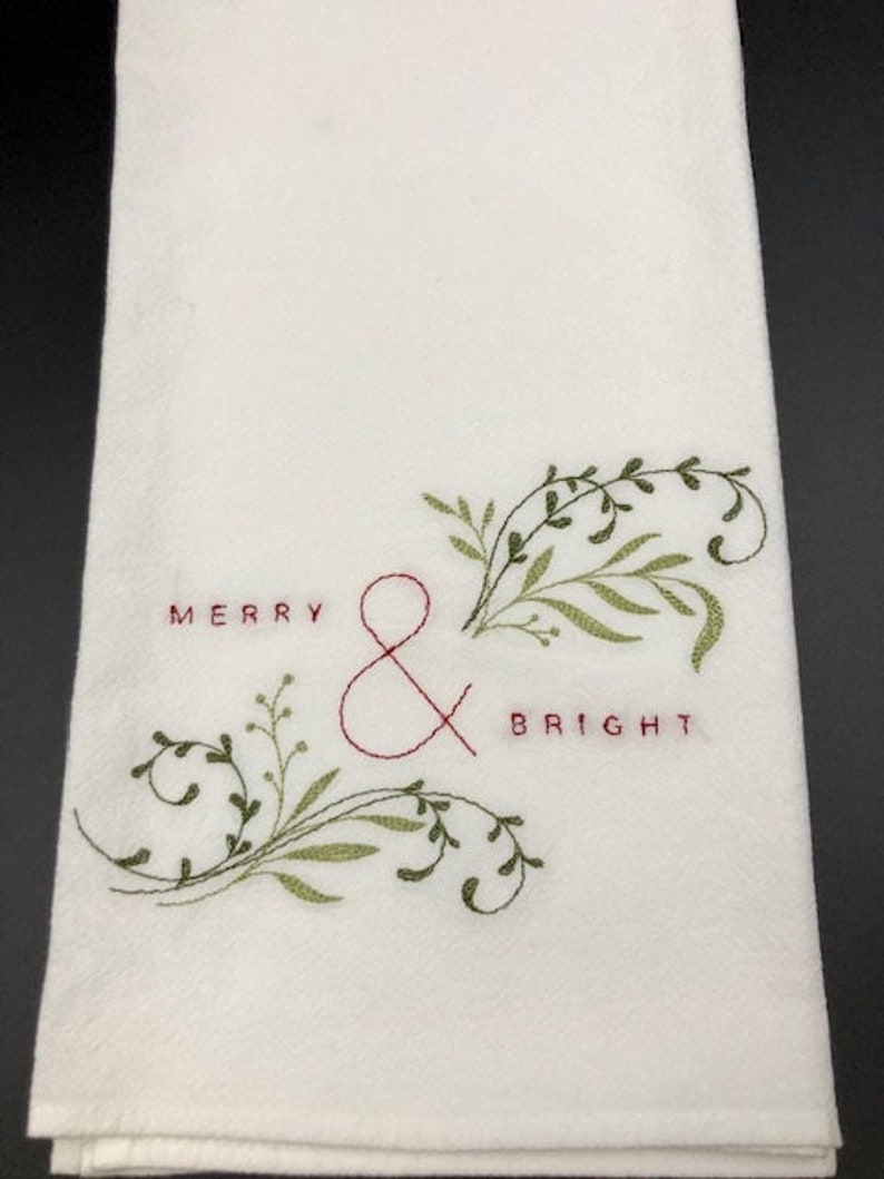 Puede incluir: Toalla de mano blanca con texto bordado y dise&ntilde;o bot&aacute;nico. Las palabras "MERRY & BRIGHT" est&aacute;n bordadas con hilo rojo. Hojas y vides verdes y oliva est&aacute;n bordadas alrededor del texto, creando un tema festivo de vacaciones.