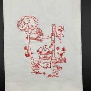 May include: Red embroidered kitchen towel with a design of a well, two geese, and a farmhouse in the background.