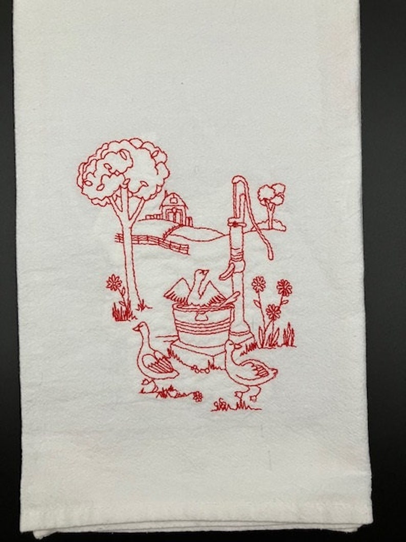 May include: Red embroidered kitchen towel with a design of a well, two geese, and a farmhouse in the background.