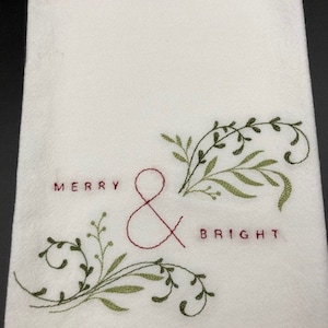 Puede incluir: Pa&ntilde;o de cocina blanco con texto bordado "MERRY & BRIGHT" en hilo rojo. Dise&ntilde;o floral bordado en verde acent&uacute;a el texto. La toalla est&aacute; hecha de una tela texturizada.
