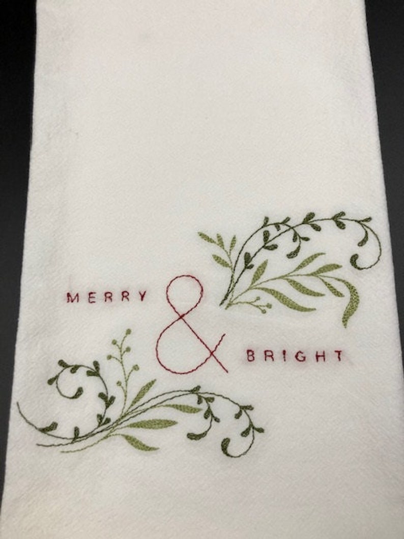 Puede incluir: Pa&ntilde;o de cocina blanco con texto bordado "MERRY & BRIGHT" en hilo rojo. Dise&ntilde;o floral bordado en verde acent&uacute;a el texto. La toalla est&aacute; hecha de una tela texturizada.