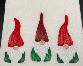 Peut inclure: Serviette de cuisine blanche avec des gnomes brodés. Chaque gnome a un chapeau rouge, une barbe blanche, un corps vert et un nez beige. Les gnomes sont disposés verticalement sur la serviette.