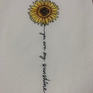 Puede incluir: Tela blanca con un diseño de girasol bordado. El girasol es amarillo con un centro marrón oscuro. Las palabras "you are my sunshine" están bordadas en escritura negra debajo de la flor.