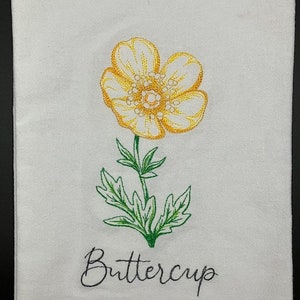 May include: White embroidered kitchen towel featuring a yellow buttercup flower with green leaves and stem. The word "Buttercup" is embroidered in cursive script below the flower. The towel is made of a soft, absorbent material.