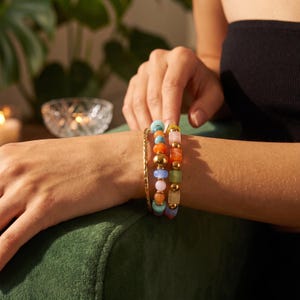 May include: A set of three bracelets worn on the wrist. One is a thin, twisted gold-tone metal band. The second features round, multi-colored beads in shades of blue, orange, pink, and gold. The third consists of square, multi-colored beads.