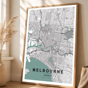 Peut inclure: Une impression encadrée d'une carte de Melbourne, Australie, dans un style minimaliste. La carte présente une palette de couleurs grise et blanche avec un accent vert d'eau pour le nom de la ville et les zones côtières. L'impression est dans un cadre en bois.