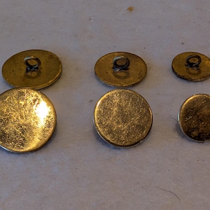May include: Six vintage brass buttons of different sizes. The buttons are round and oval, with a loop on the back for sewing. The brass has a worn, aged appearance, with a golden hue.