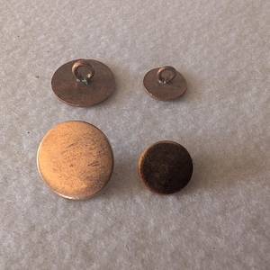 Op de afbeelding: Vier antieke koperen knopen in verschillende maten. Twee knopen hebben een lus aan de achterkant, de andere zijn plat. De knopen vertonen tekenen van ouderdom, met zichtbare textuur- en kleurvariaties.