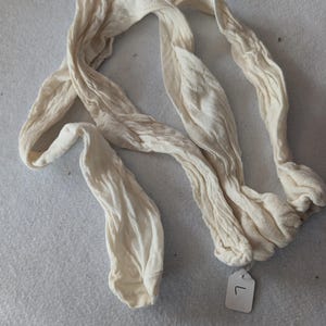 May include: Cream-coloured, textured fabric garment, possibly stockings or tights, arranged on a light grey surface. The fabric is bunched and wrinkled, with a tag labelled "L" attached. The item appears to be vintage or handmade.