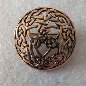Op de afbeelding: Een ronde, bronskleurige broche met een ingewikkeld Keltisch knoopontwerp. De broche heeft verweven lijnen die een doorlopend patroon vormen. Aan de achterkant is een speld bevestigd om de broche aan kleding of accessoires te bevestigen.