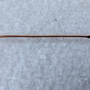 Könnte beinhalten: Eine schlanke, kupferfarbene Nadel mit einer scharfen Spitze und einem kleinen, runden Öhr. Die Nadel ist gerade und scheint aus Metall zu sein. Sie liegt auf einer hellgrauen, strukturierten Oberfläche. Ca. 4 cm lang.