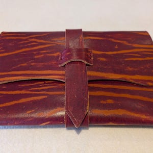 May include: A burgundy leather clutch with orange-brown streaks and a matching strap closure. The rectangular wallet-style bag has a flap closure and a vertical strap that secures the front. The leather has a natural, textured appearance.