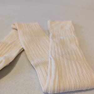 May include: A pair of cream-coloured, ribbed knit leg warmers. The leg warmers are folded and lying on a light-coloured surface. The ribbed texture is visible, and the fabric appears soft and comfortable. These leg warmers are a cosy accessory.