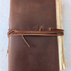 May include: A brown leather journal with a leather tie wrapped around it. The pages are visible on the right, with a rough, textured edge. The leather has a worn appearance.