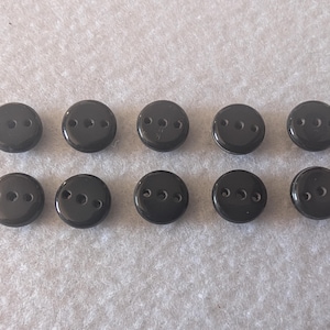 May include: Ten glossy, black, round buttons with three holes each are arranged in two rows. These buttons are likely for sewing or crafting projects. The buttons are evenly spaced and appear to be made of a hard, durable material.