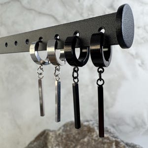 May include: A display of hoop earrings with dangling bar pendants. The earrings are silver and black, hanging from a black leather display. The earrings are displayed against a neutral background.
